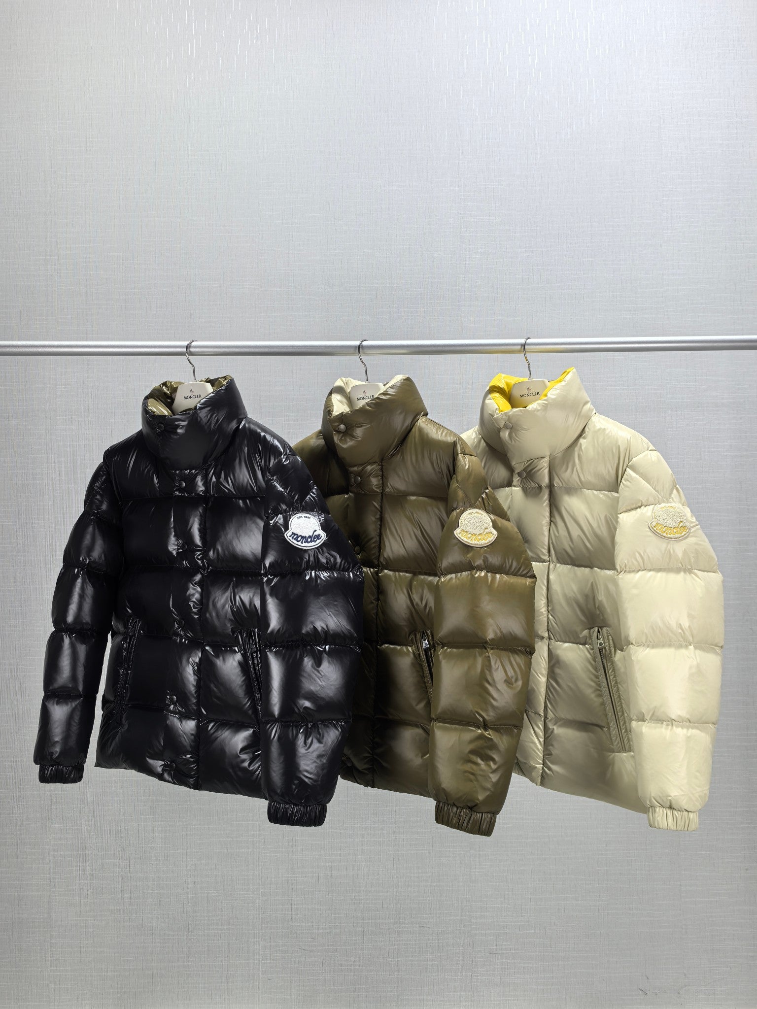 Three Moncler puffer jackets on hangers: black, olive, and light, front view