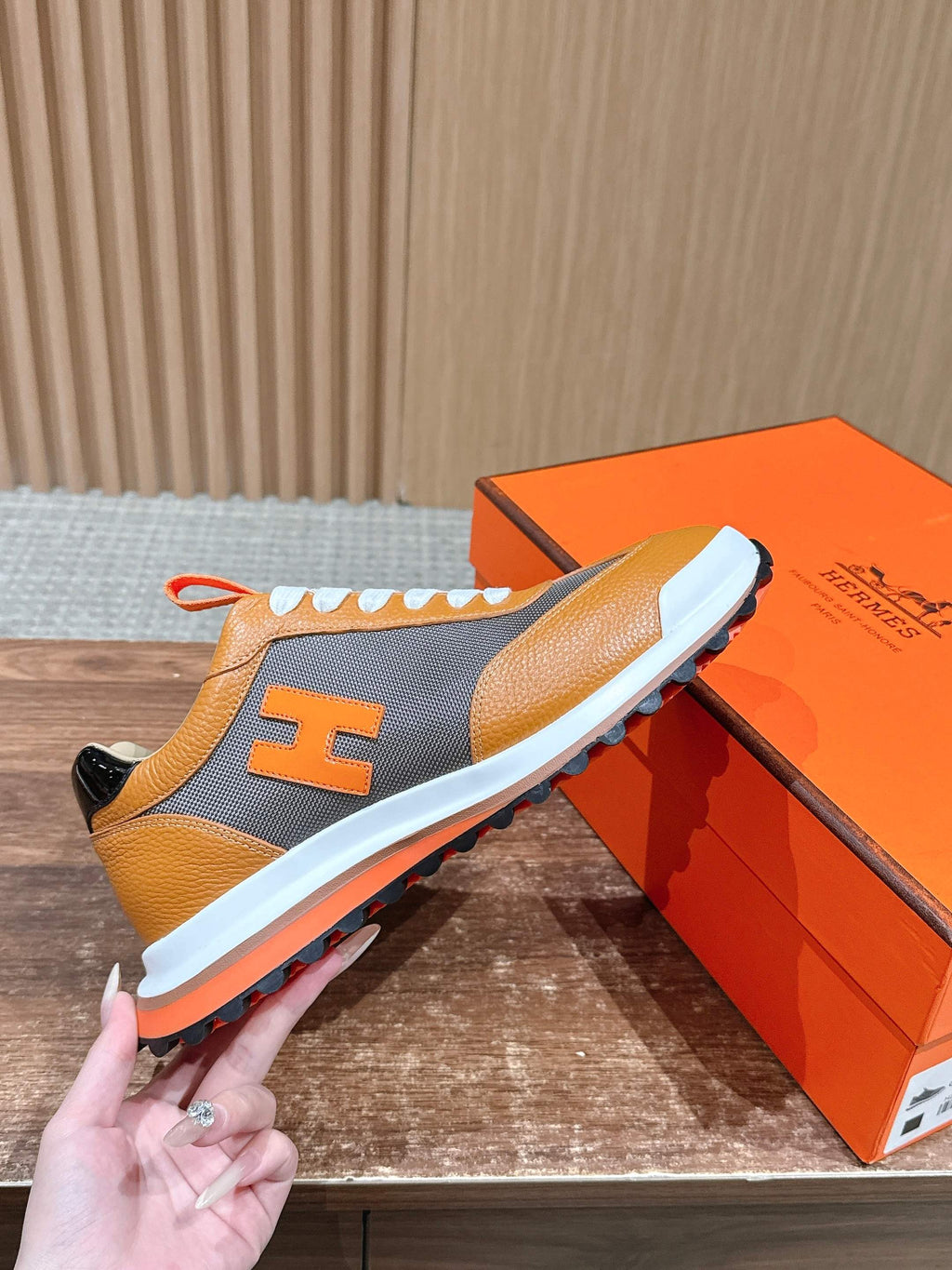 Hermes sneaker held by hand, orange leather, gray side panels, white laces