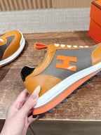 Hermes sneaker held front-side, orange and gray upper with white laces and orange trim