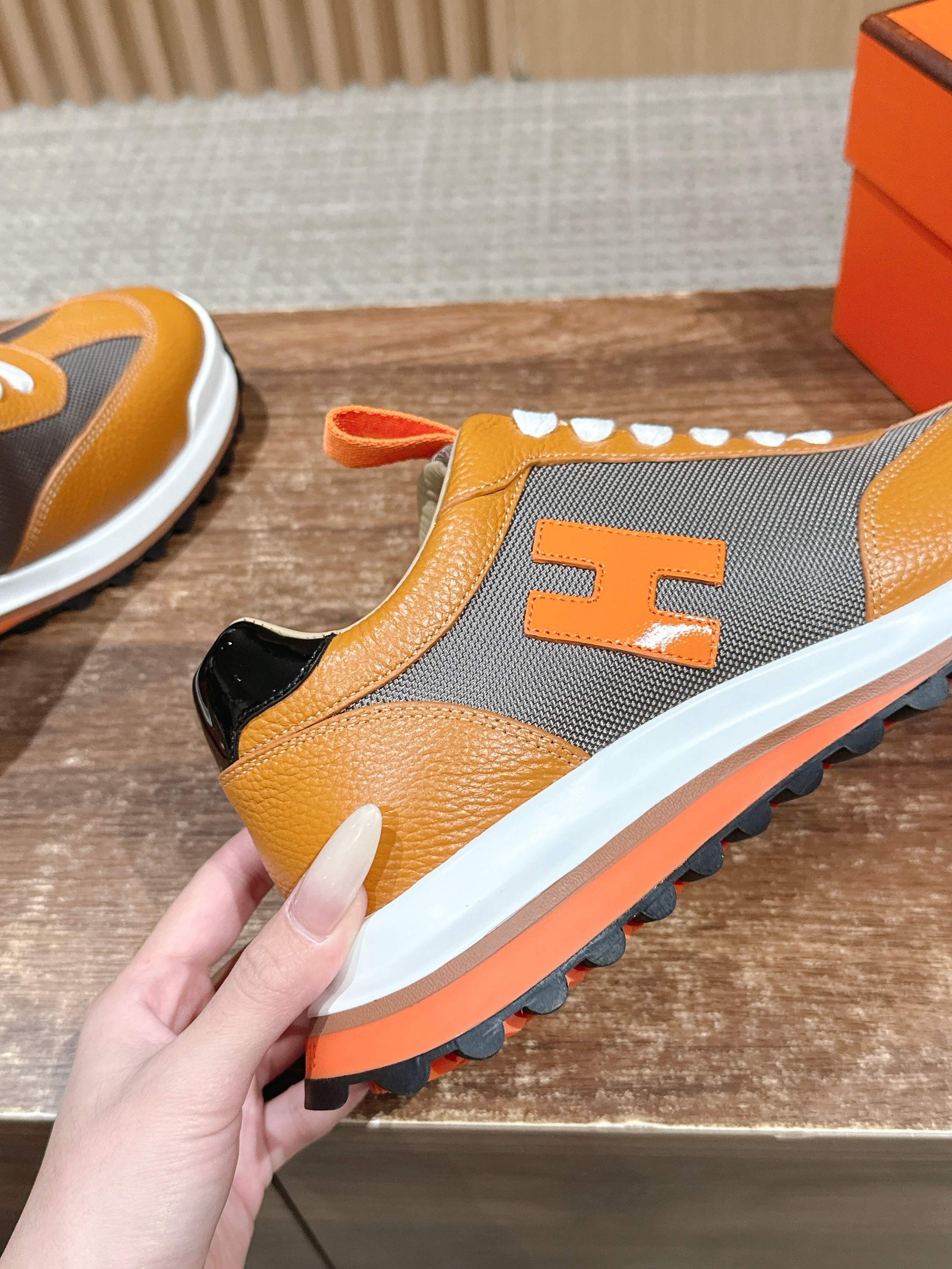 Hermes sneaker held front-side, orange and gray upper with white laces and orange trim