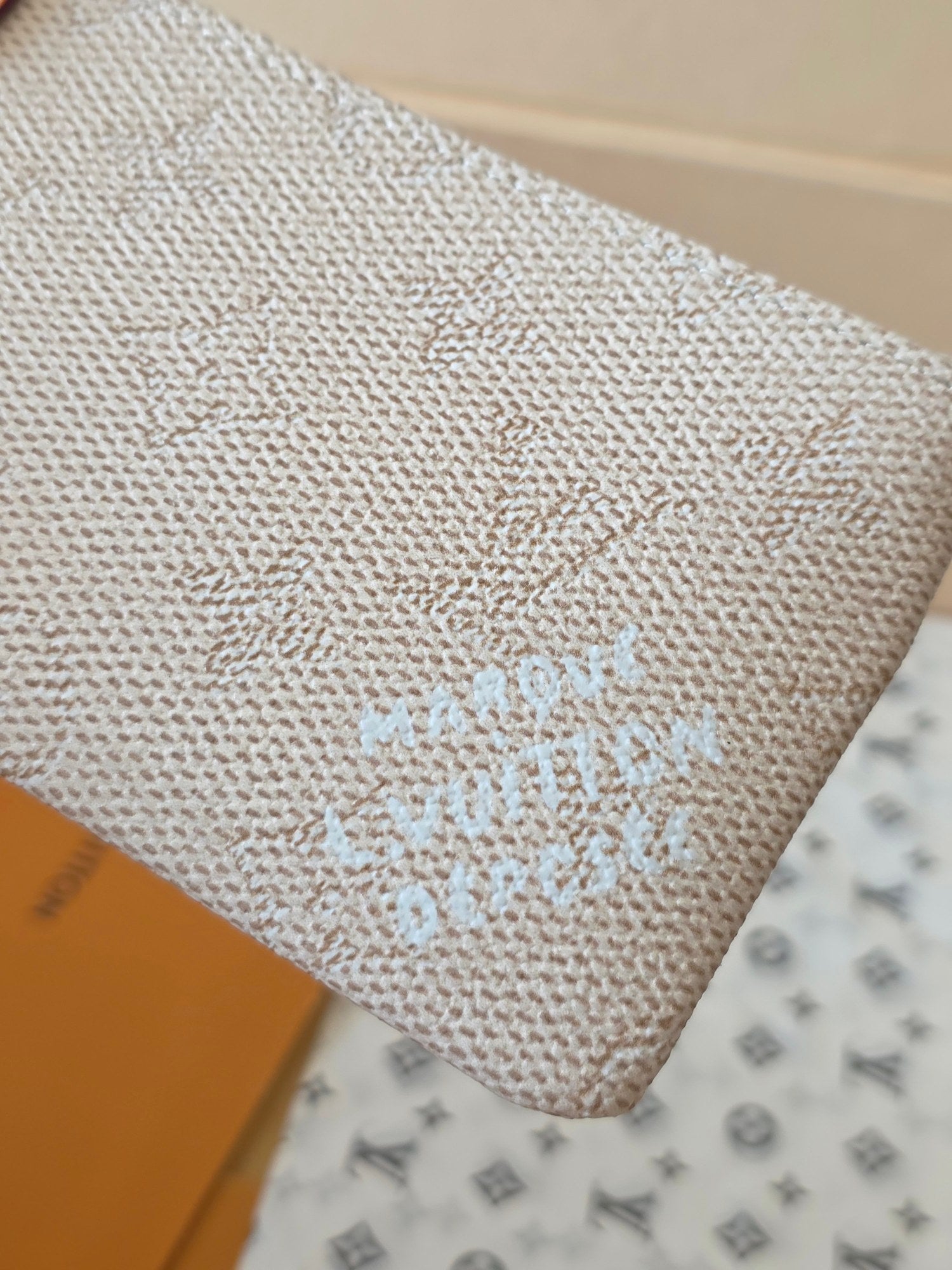 Close-up of light beige textured pouch front with faint monogram logo