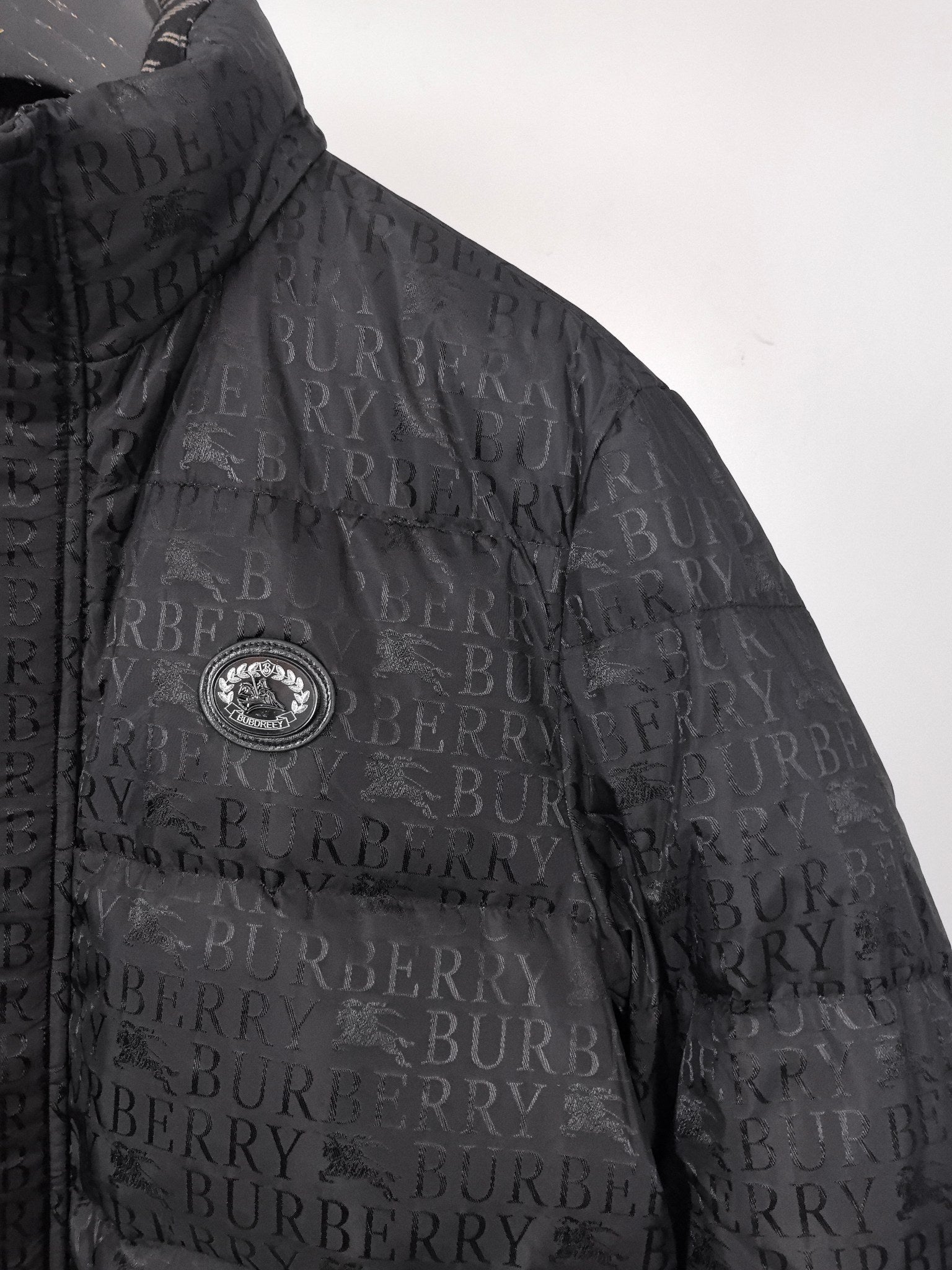 Long black puffer jacket with circular chest emblem and repeating Burberry pattern