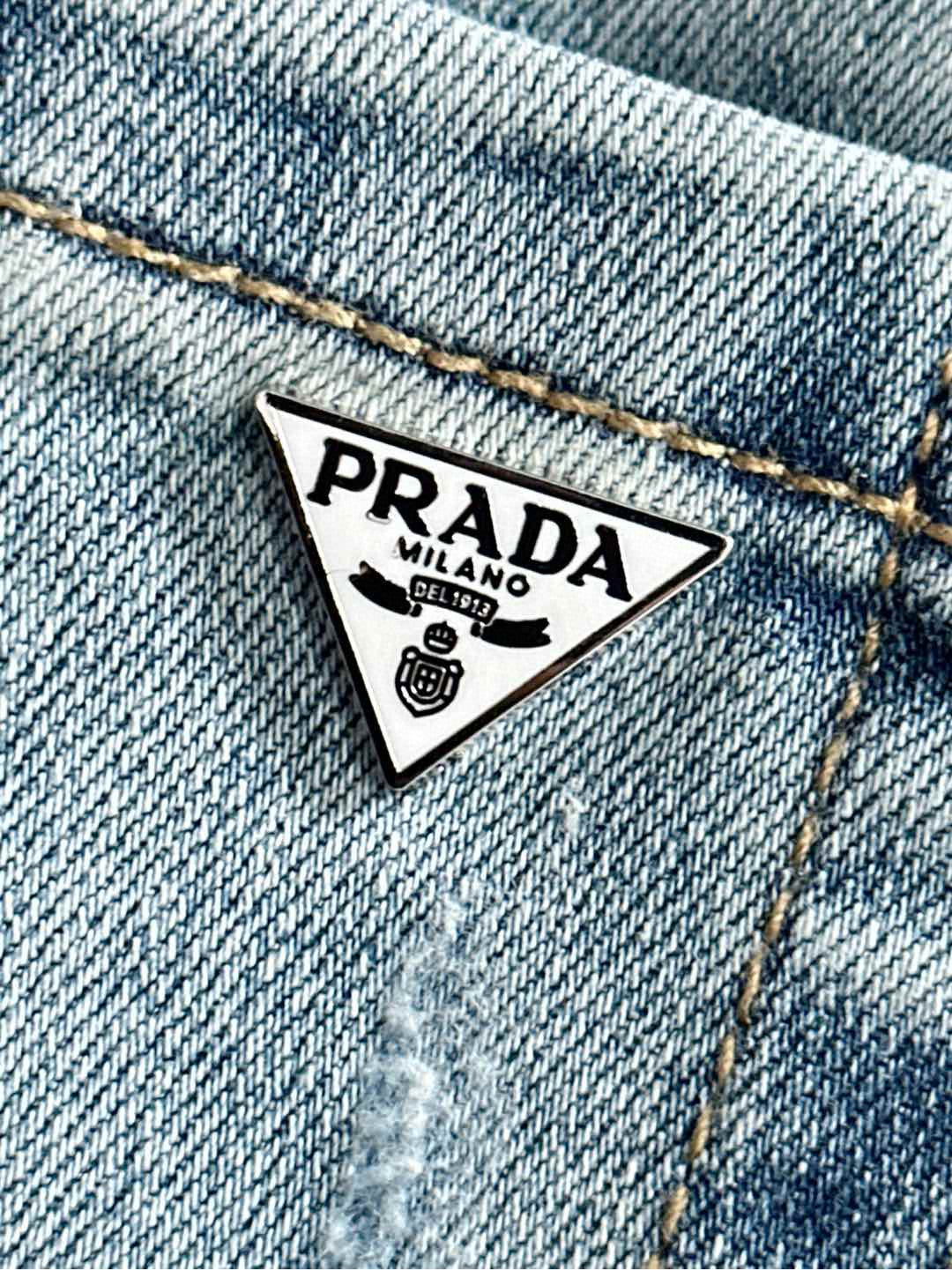 PRADA triangle logo patch on light blue denim pocket, stitched with gold thread.