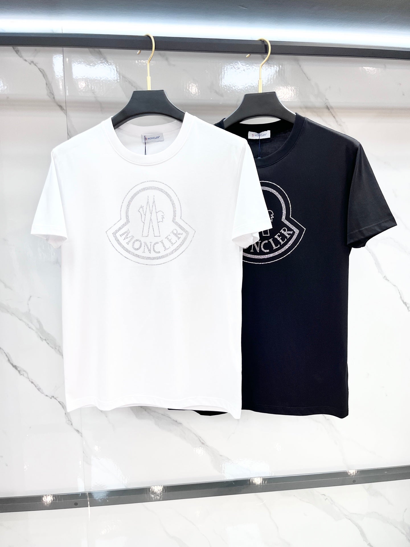 White and black Moncler T-shirts with circular chest logos on hangers.