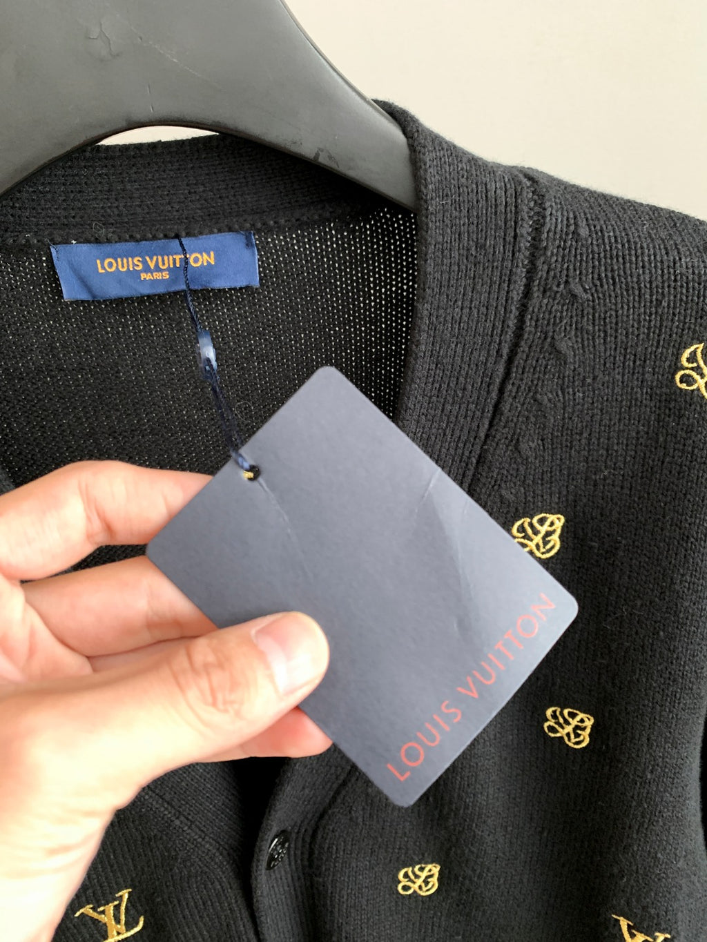 Hand holding Louis Vuitton tag on dark knit cardigan with gold monogram embroidery