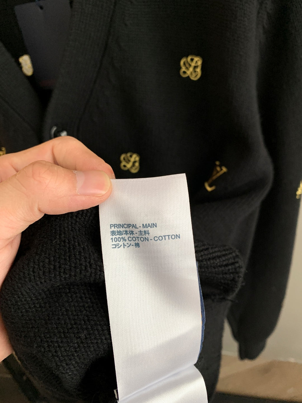 Fabric content tag held up: 100% cotton, on dark knit cardigan with gold pattern