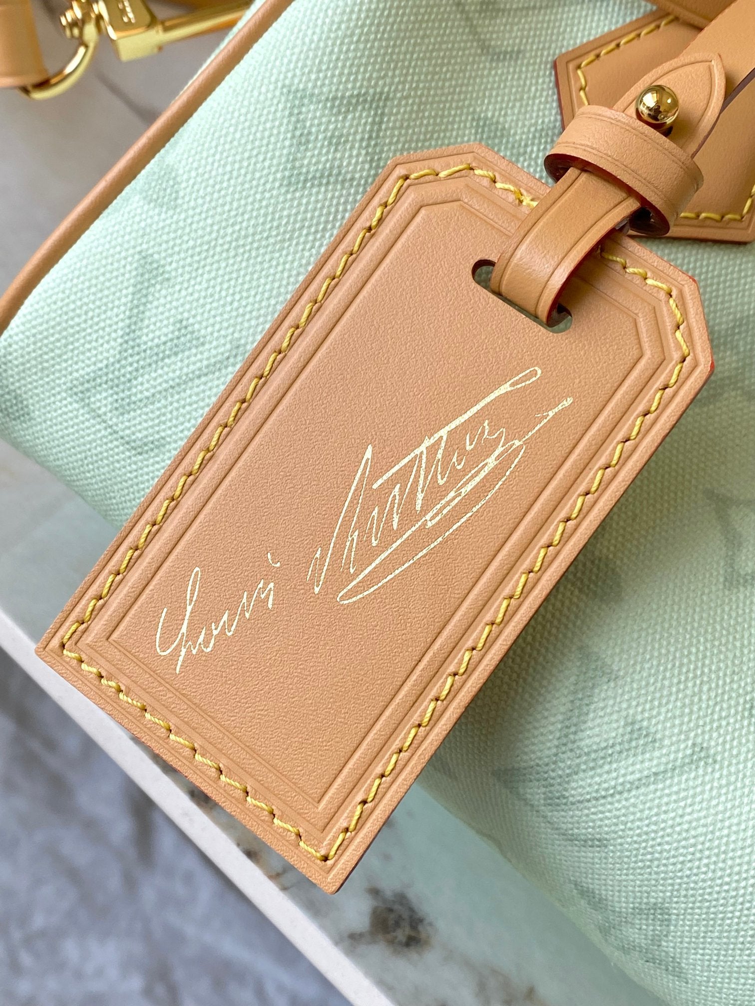Mint green handbag with tan leather name tag, gold trim, and embossed cursive text