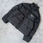 Long puffer jacket in dark gray, laid flat, with visible hood and logo patch