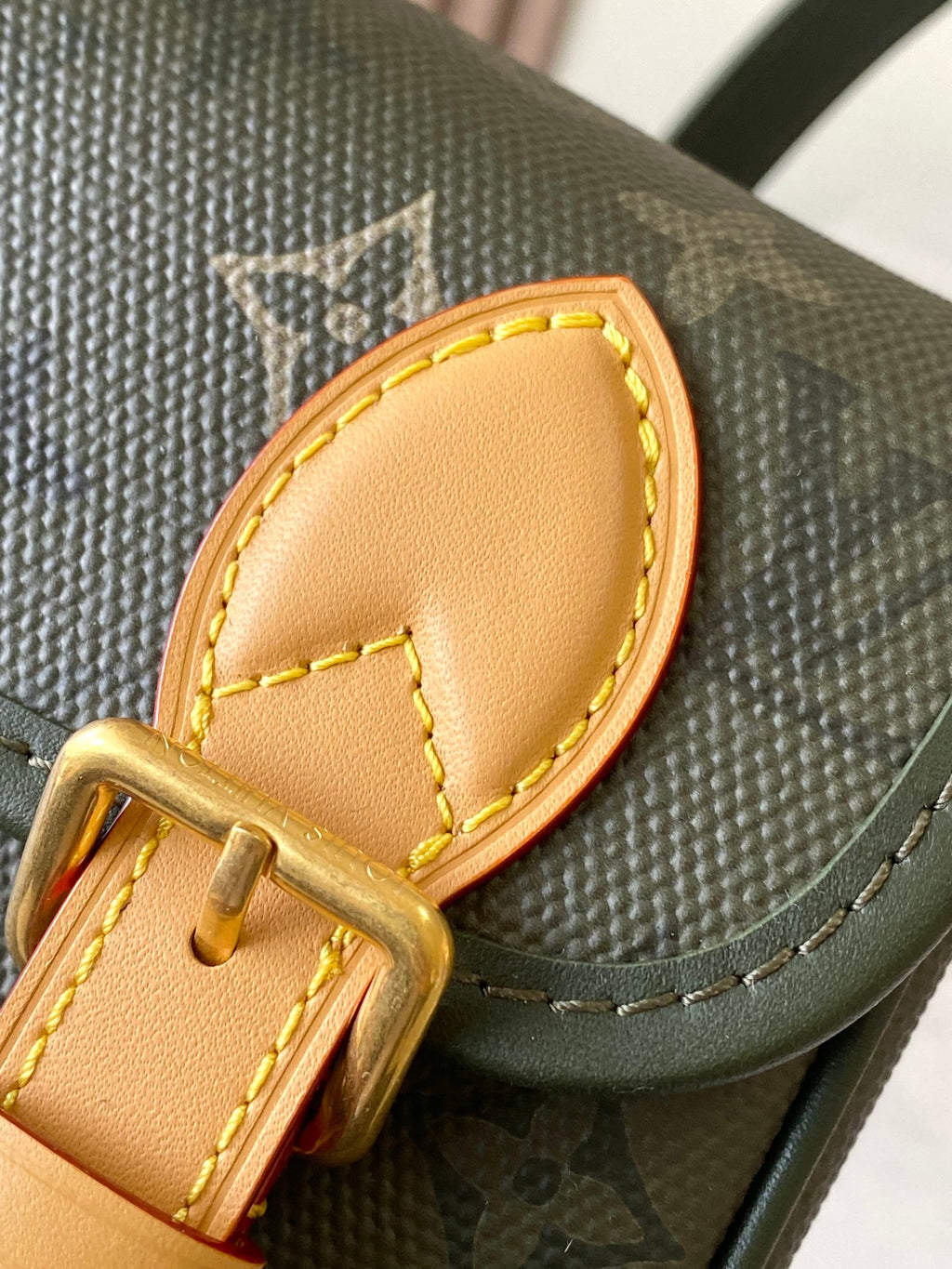 Close-up of tan leather trim and gold buckle on dark green Louis Vuitton monogram bag.