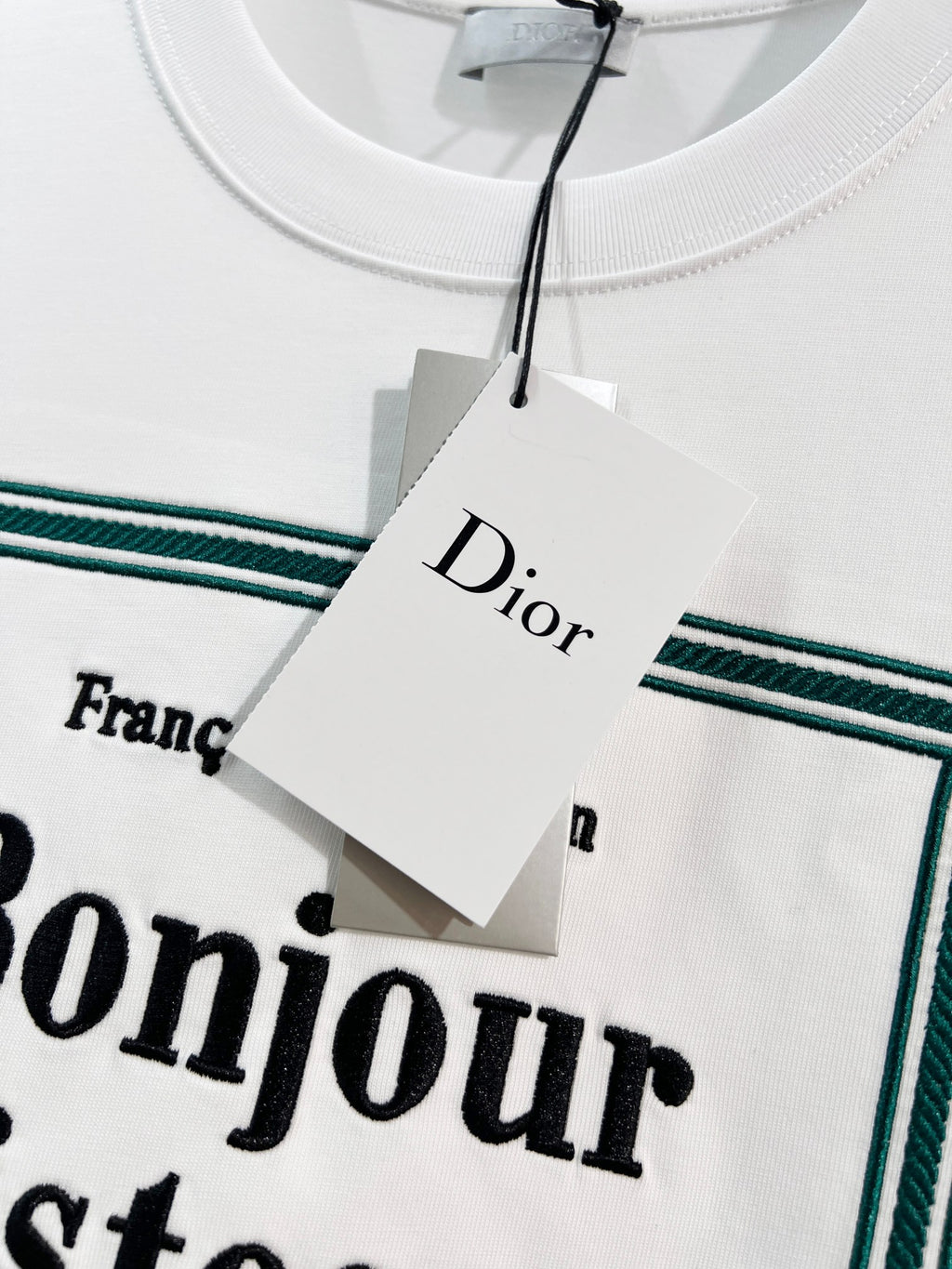 White Dior T-shirt neckline and hanging tag over green-bordered “Bonjour tristesse” print