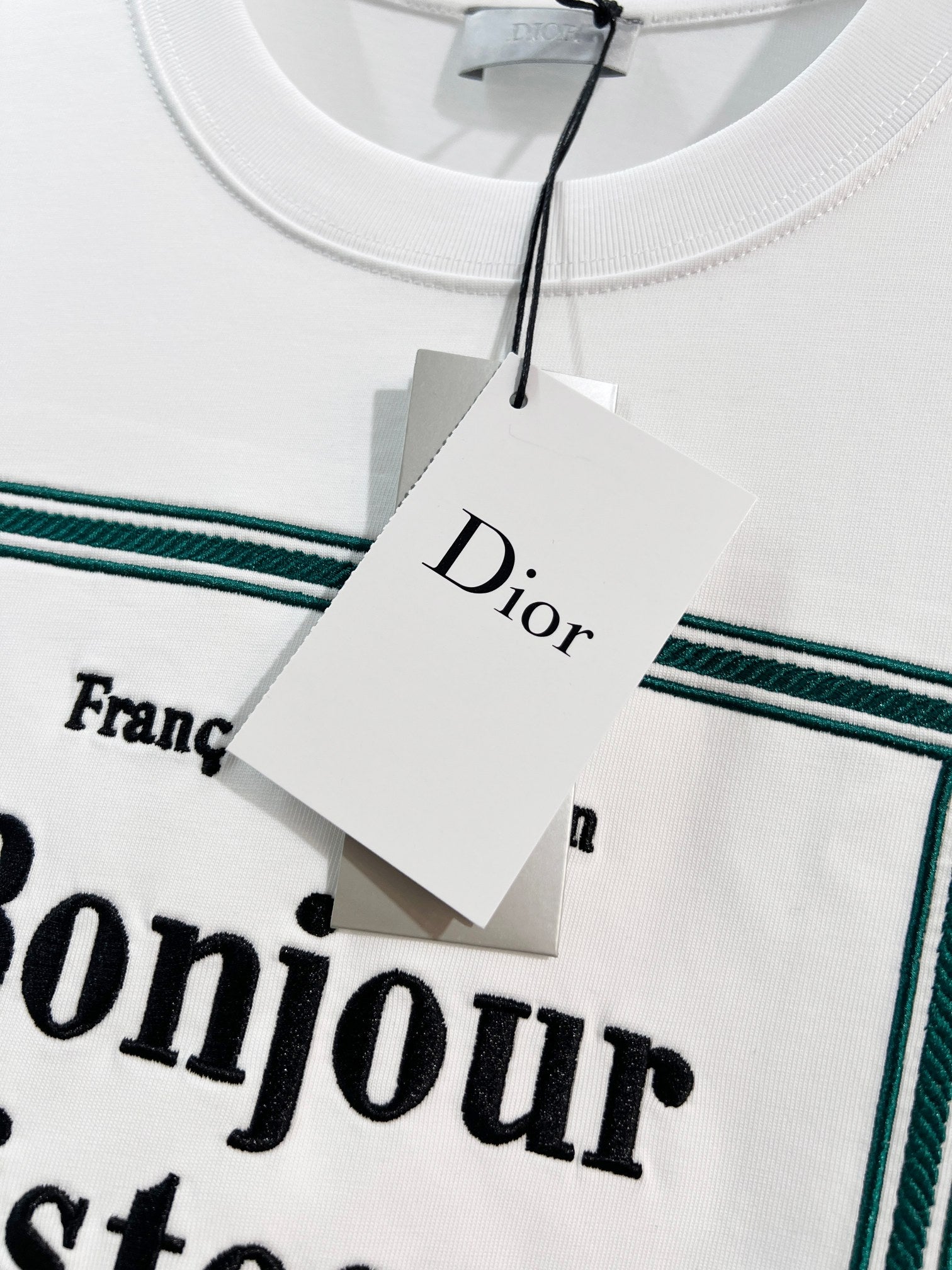 White Dior T-shirt neckline and hanging tag over green-bordered “Bonjour tristesse” print