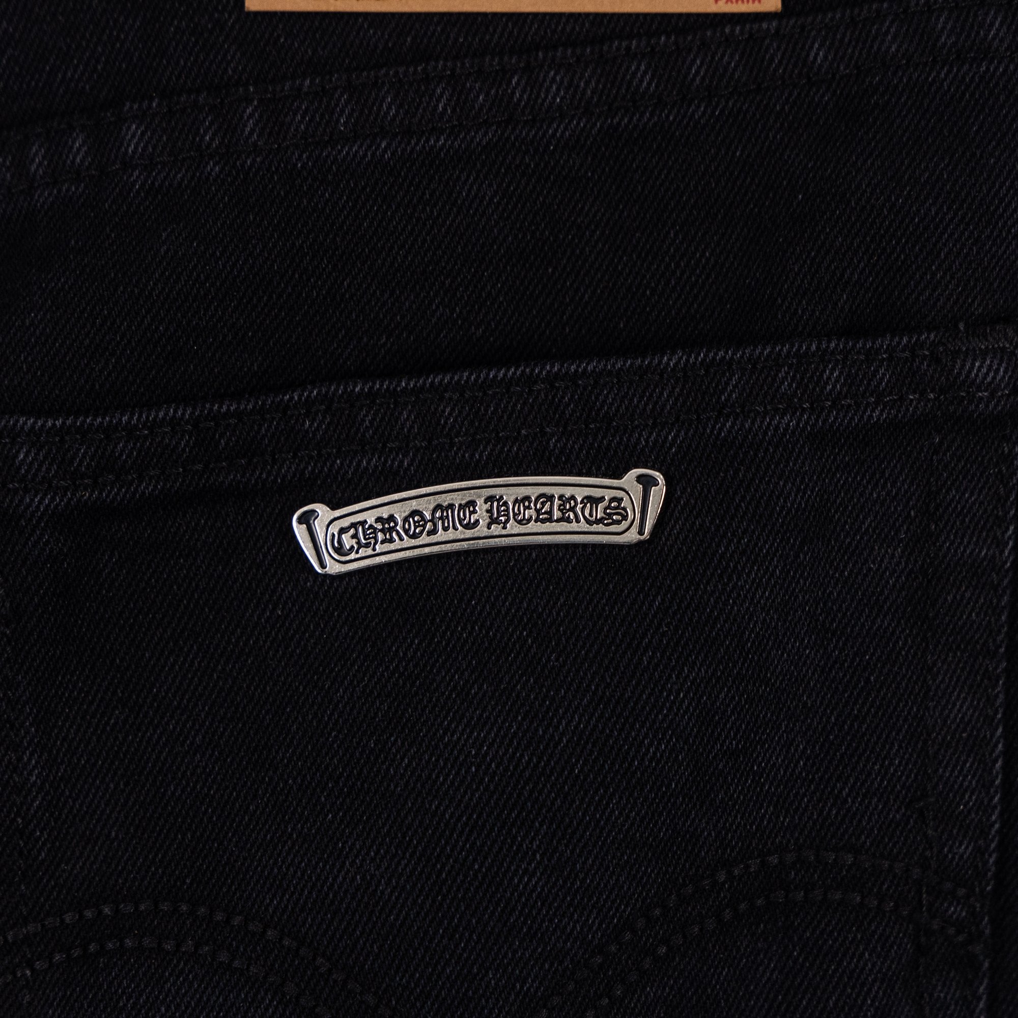 Metal logo patch on dark denim pocket seam, small rectangular tag.