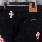 Dark denim pants front with pink cross patch, metal button and branded waistband label.