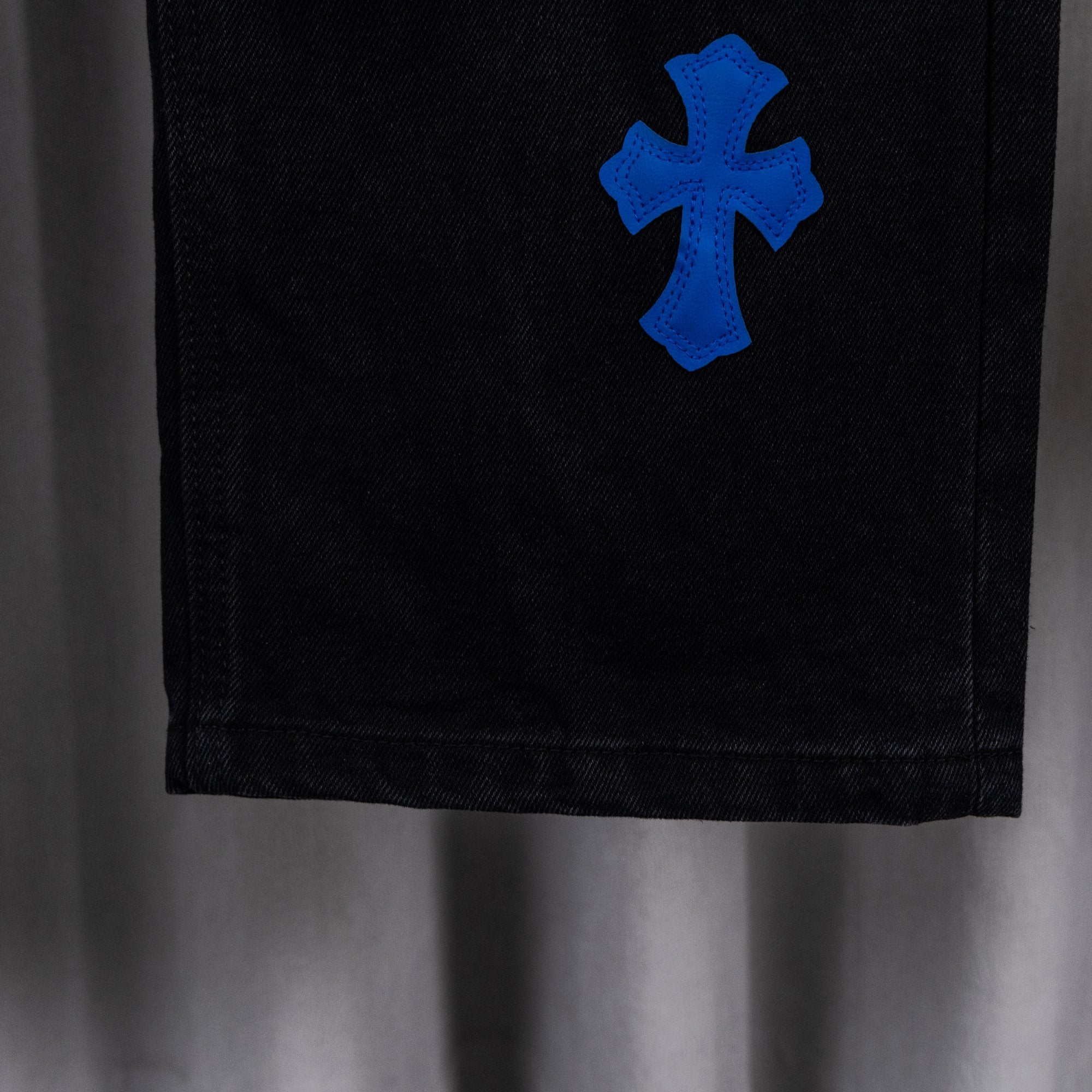 Black denim pant leg with blue cross applique, cropped close-up view.