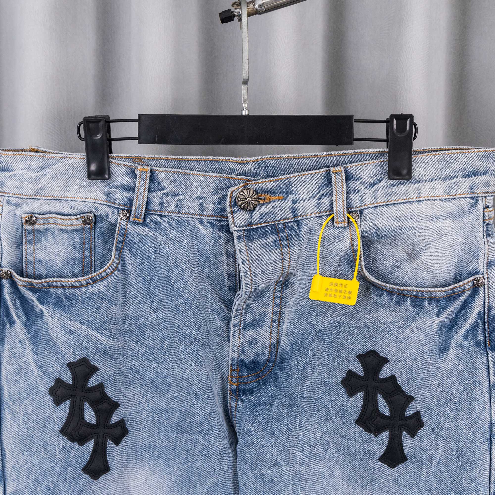 Light blue denim jeans with black cross patches, waistband pockets, and yellow tag