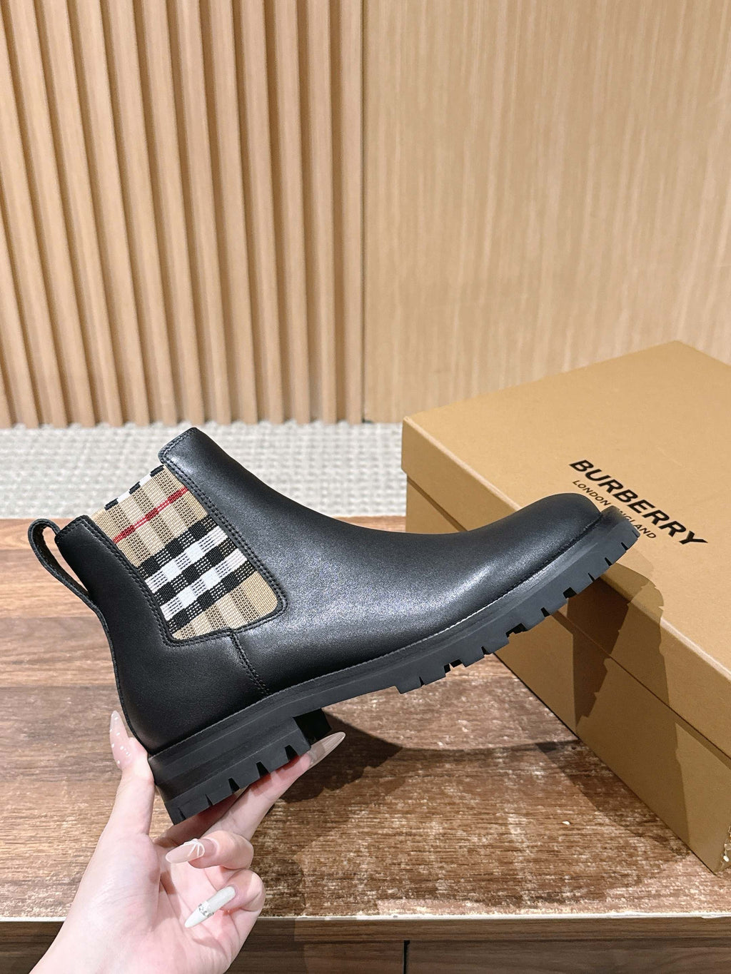 Black Chelsea boots with check-pattern elastic panel, held next to box