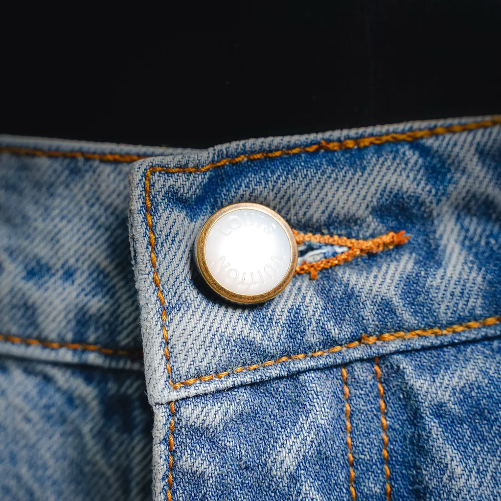 Round gold-tone button on light blue denim pocket with orange contrast stitching