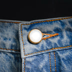 Round gold-tone button on light blue denim pocket with orange contrast stitching