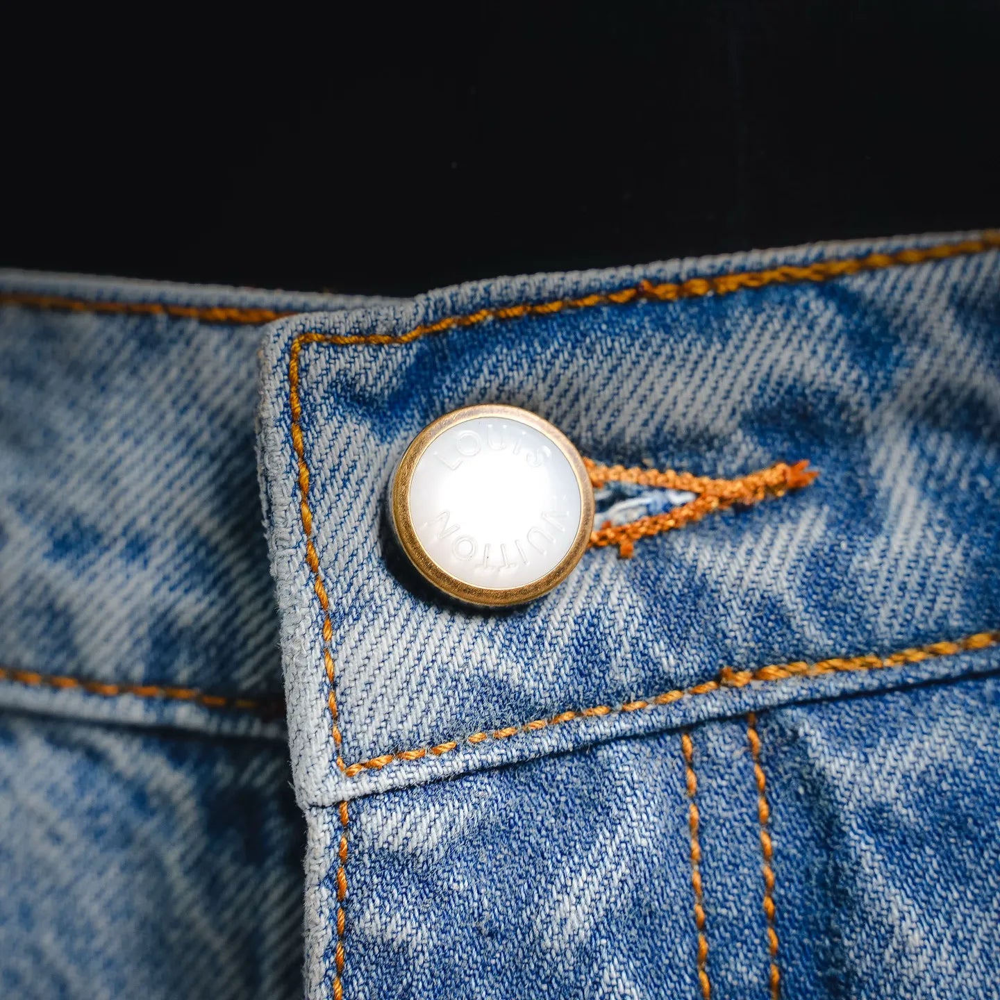 Round gold-tone button on light blue denim pocket with orange contrast stitching
