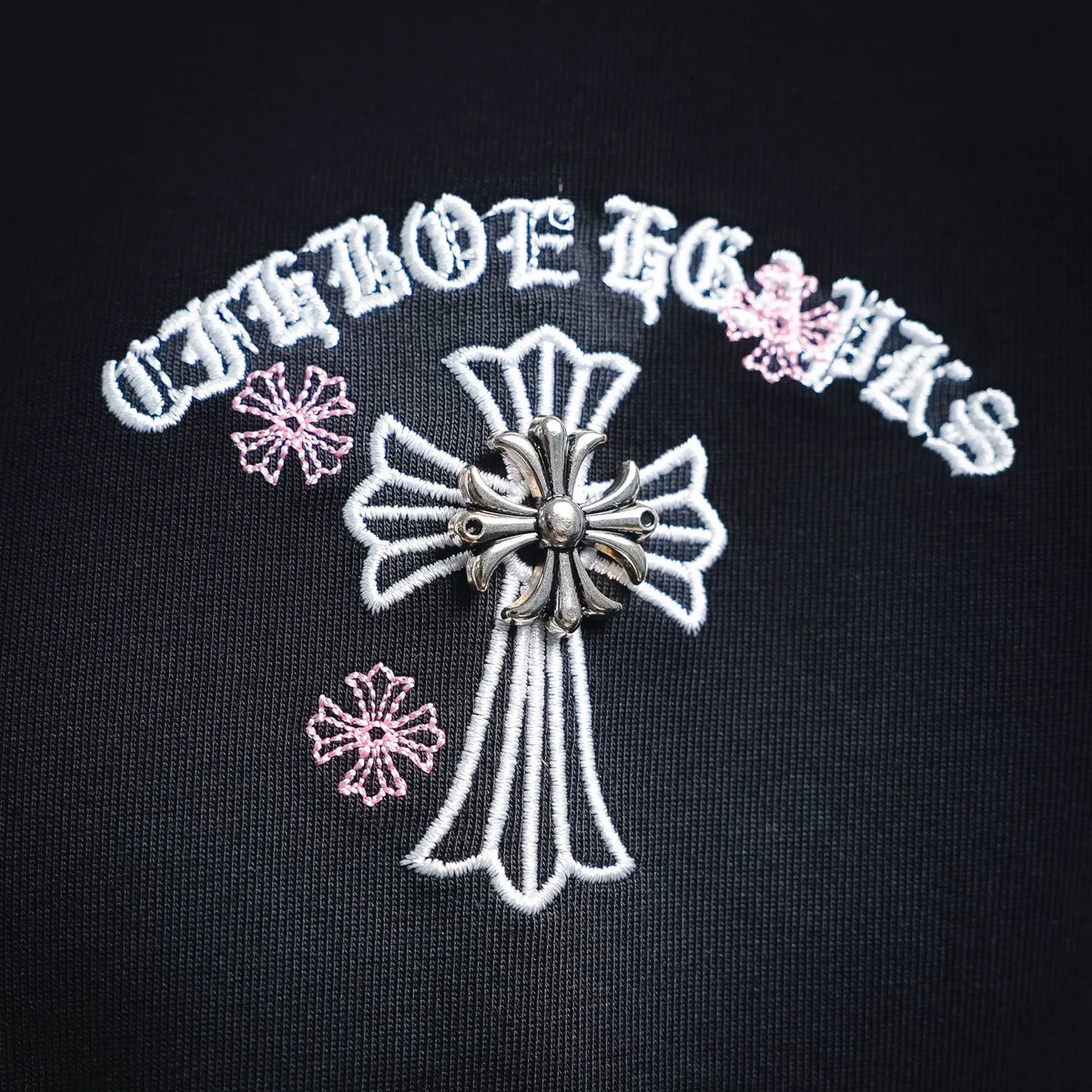Dark T-shirt front cross emblem with outlined chain details and pink flower accents.