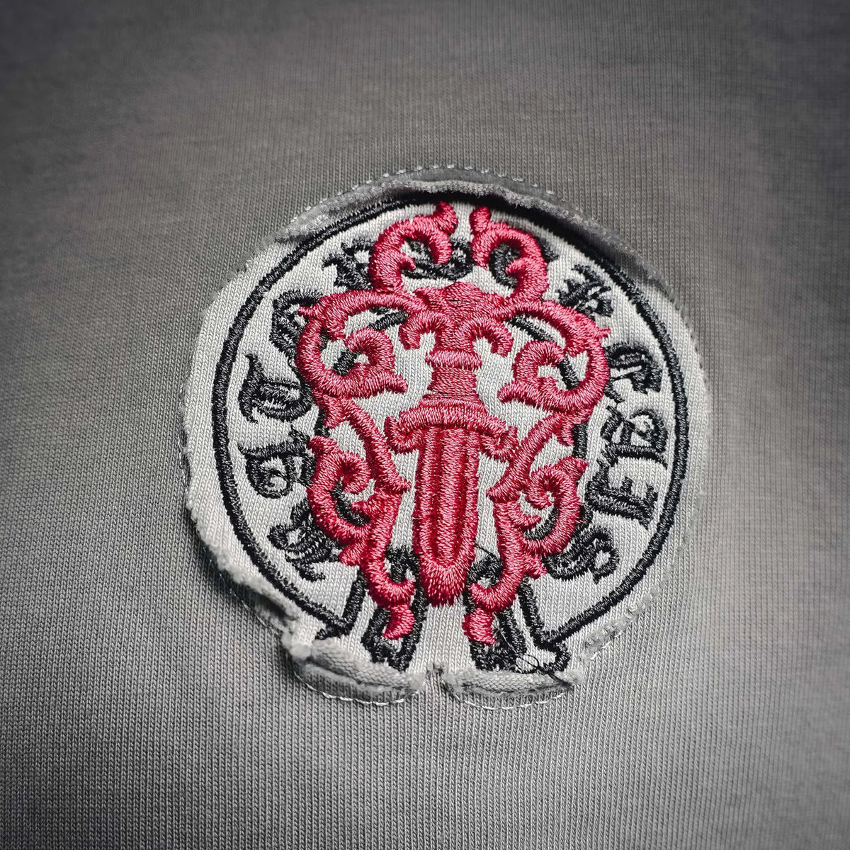 Embroidered round crest patch with red emblem and dark text on gray T-shirt.