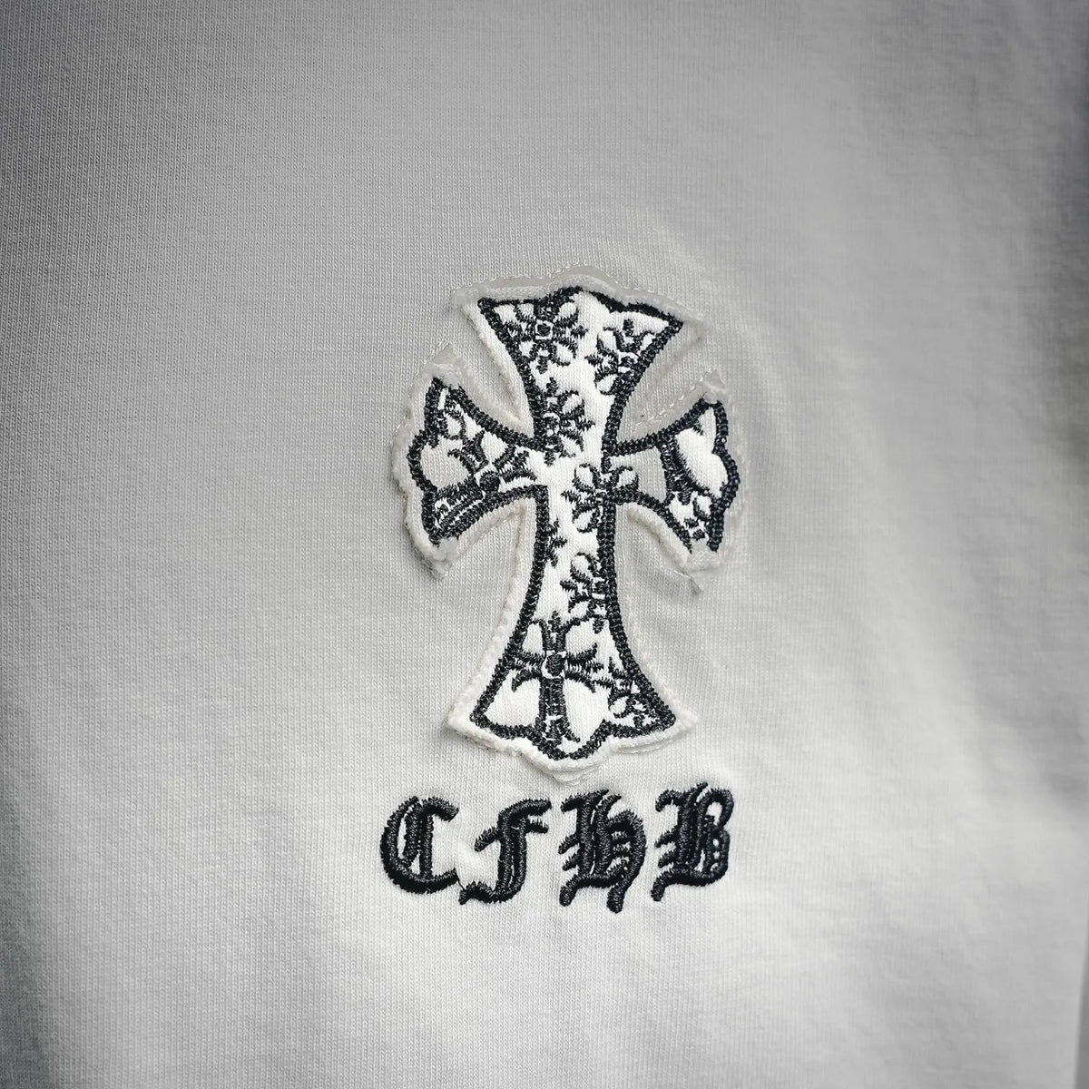 White T-shirt with detailed black cross patch and stylized letters below.