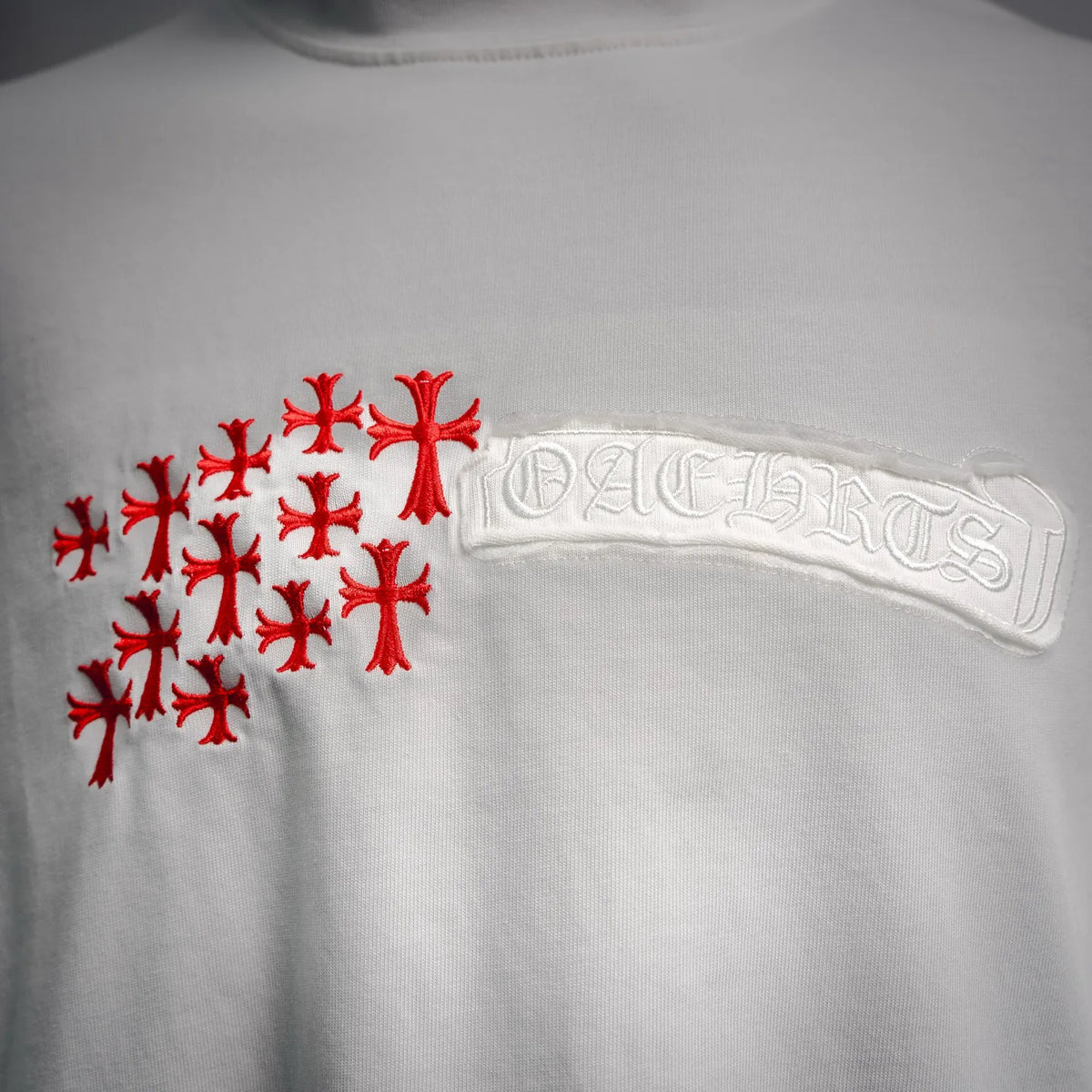 White T-shirt front close-up: red cross pattern beside embossed logo patch