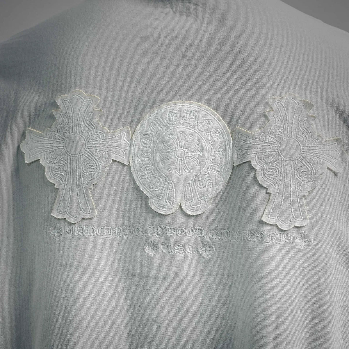Raised embossed-style graphic on white T-shirt back: emblem with cross details