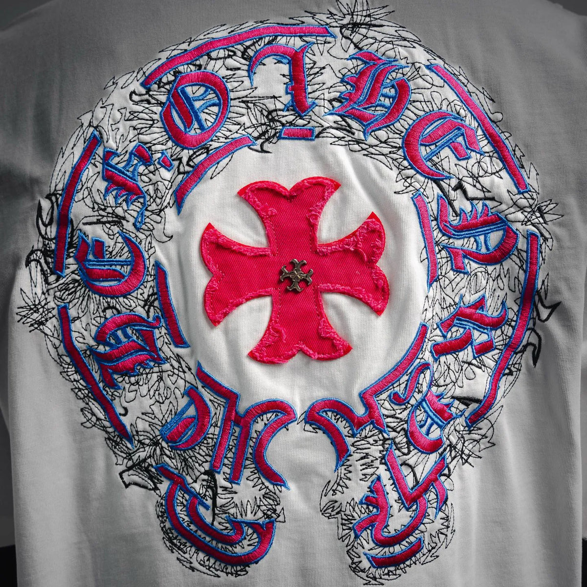 White T-shirt back with circular embroidered emblem featuring red flower and blue accents.