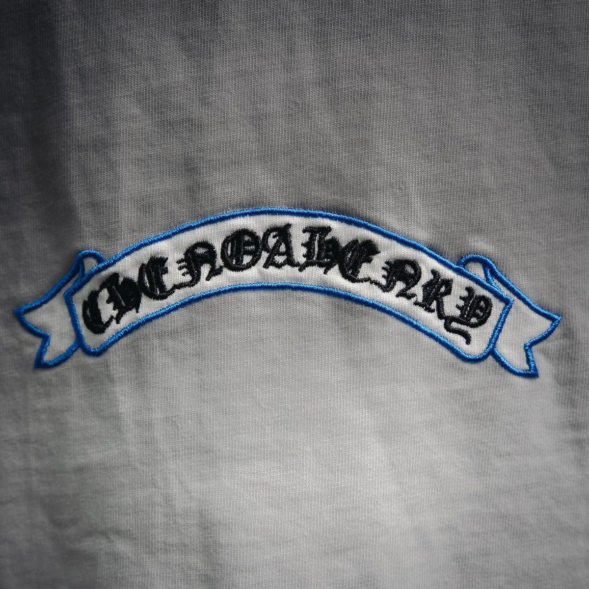 White T-shirt sleeve patch text embroidery in black with a blue border.