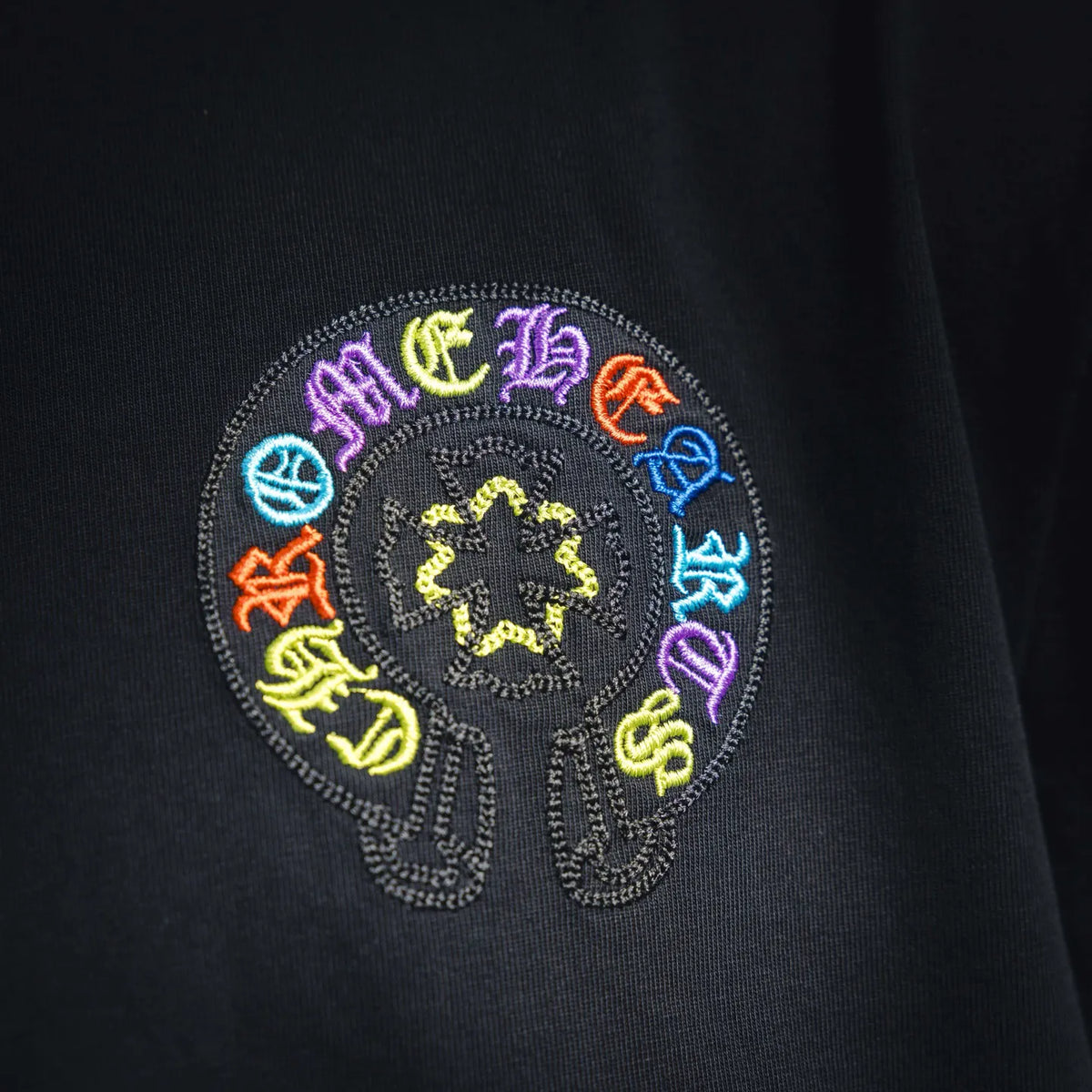 Embroidered circular graphic on black T-shirt chest with rainbow-colored text.