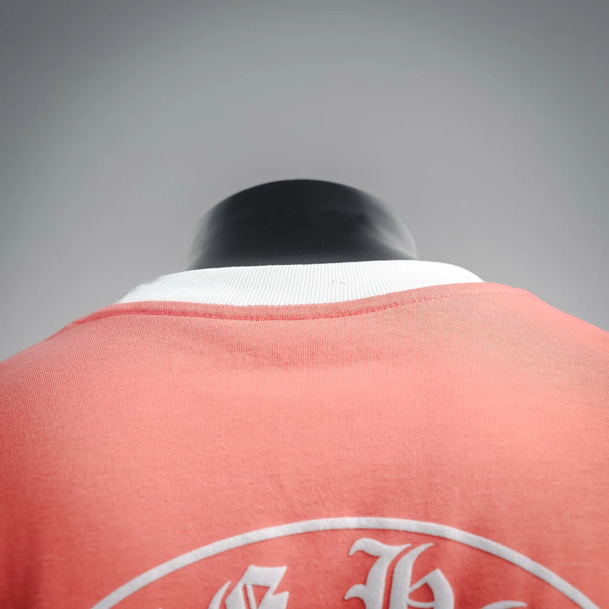 Coral pink T-shirt back view with white collar trim and partial circular emblem graphic