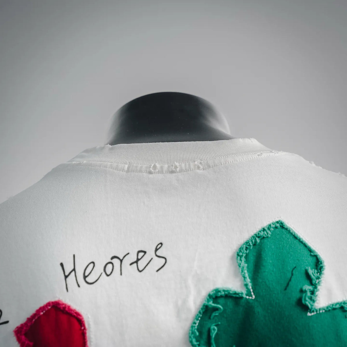 White T-shirt with 'Heor's' text and green embroidered flower patch at upper back
