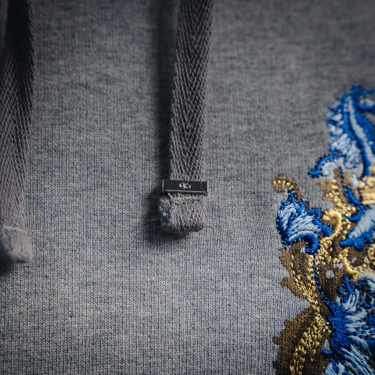 Gray hoodie neckline with small black brand tag and detailed embroidered crest artwork