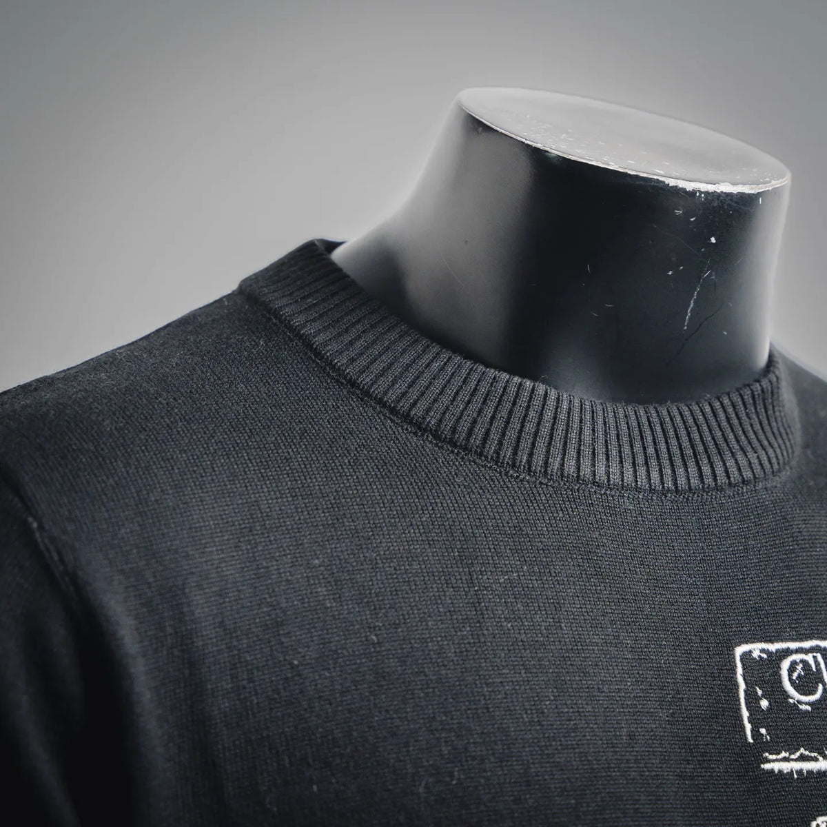 Black crewneck knit sweater with ribbed collar and white logo patch on chest