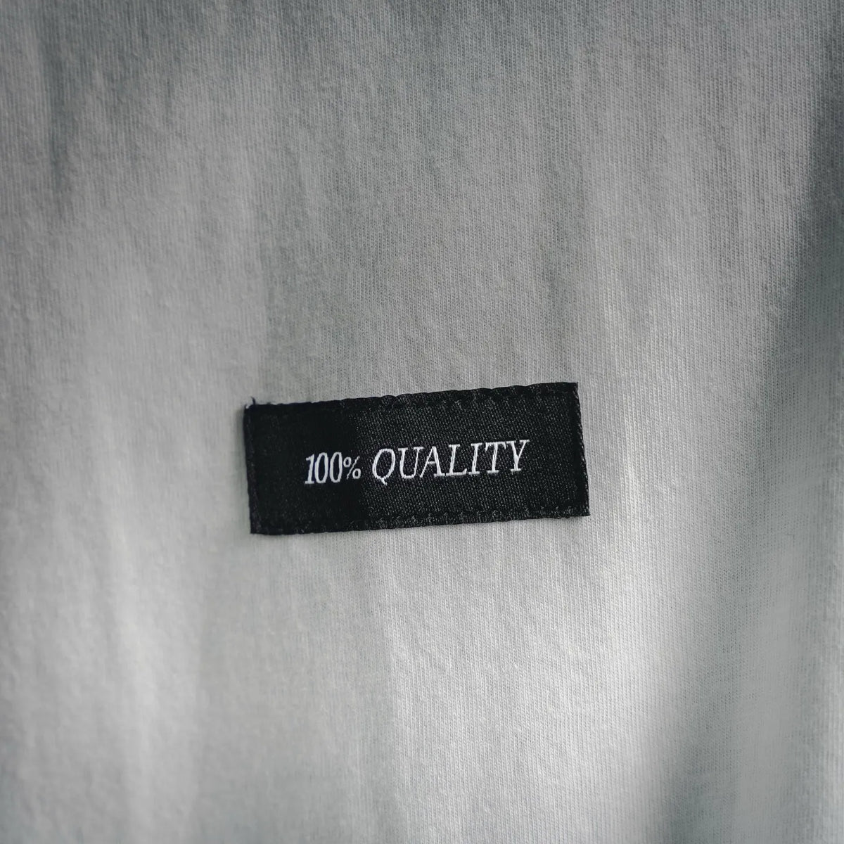 Black fabric label reading '100% QUALITY' on white T-shirt