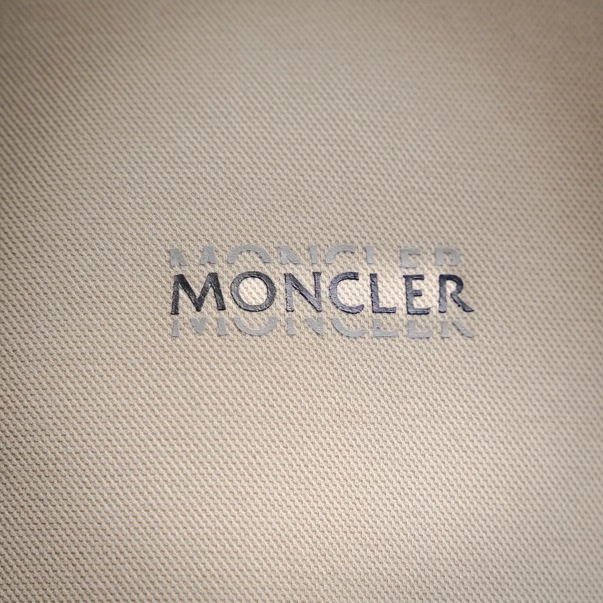 Moncler logo on light beige textured polo fabric, printed in dark lettering.