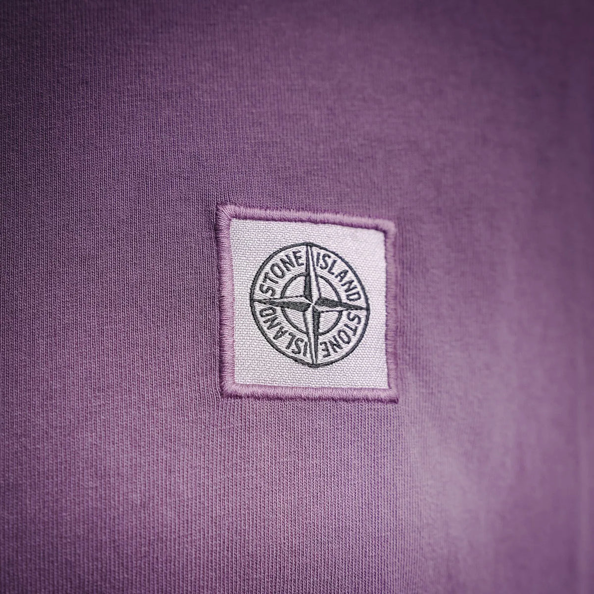 Stone Island logo patch on purple T-shirt chest, rectangular embroidered badge