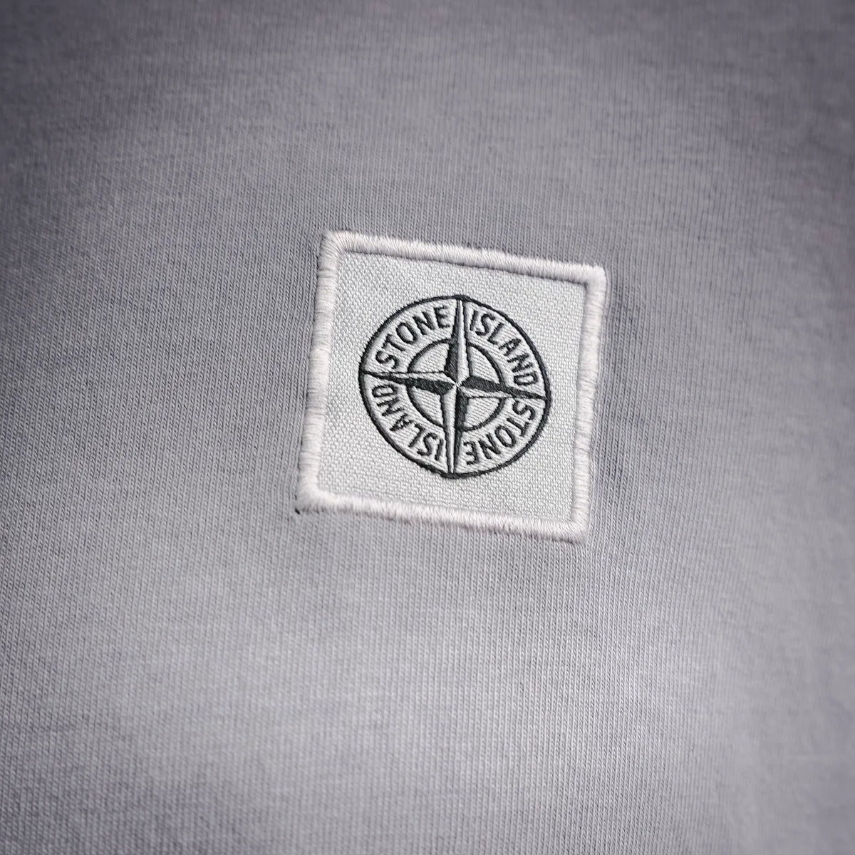 Light gray Stone Island T-shirt with embroidered chest logo patch in white square.