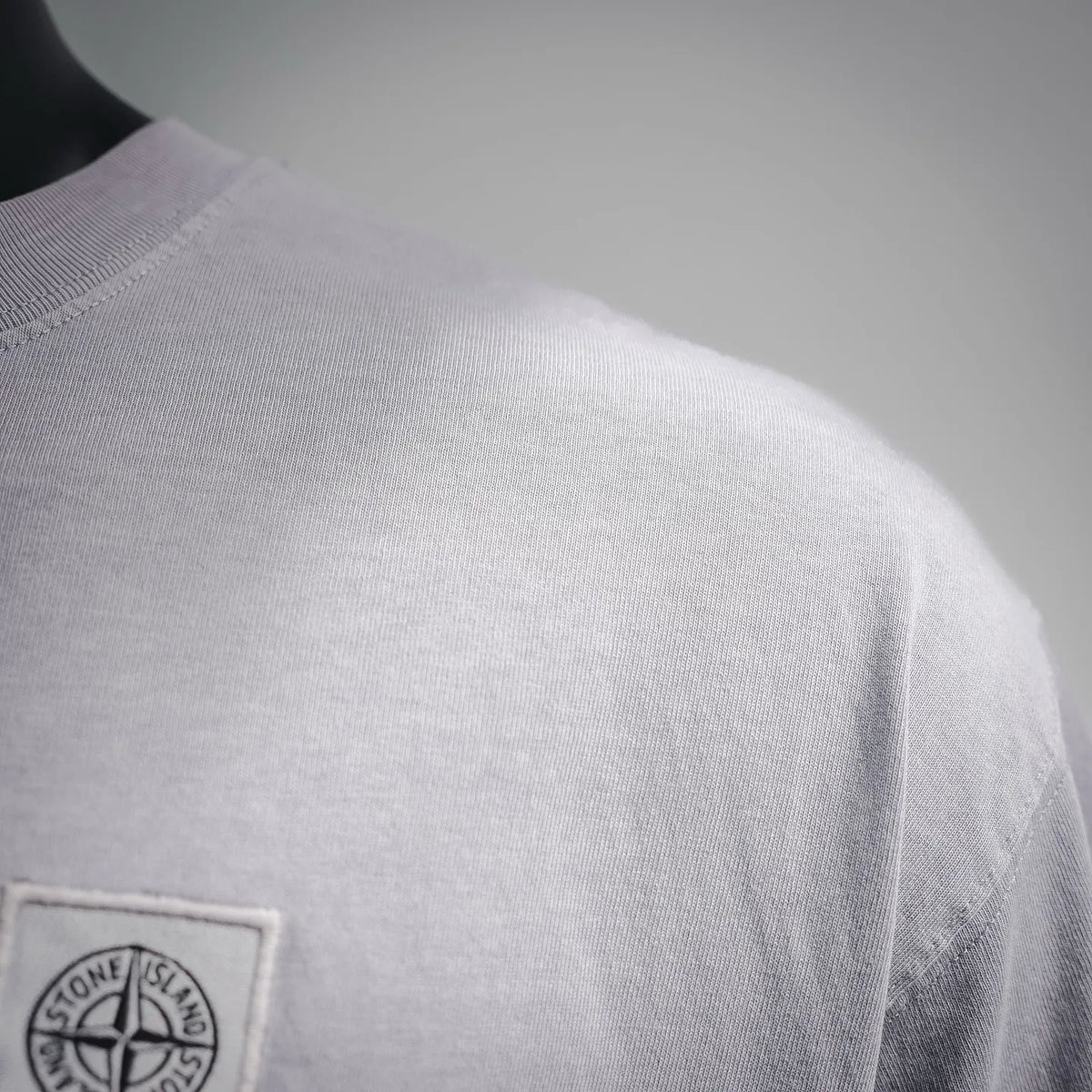 Light gray Stone Island T-shirt with chest logo patch, close-up at shoulder.