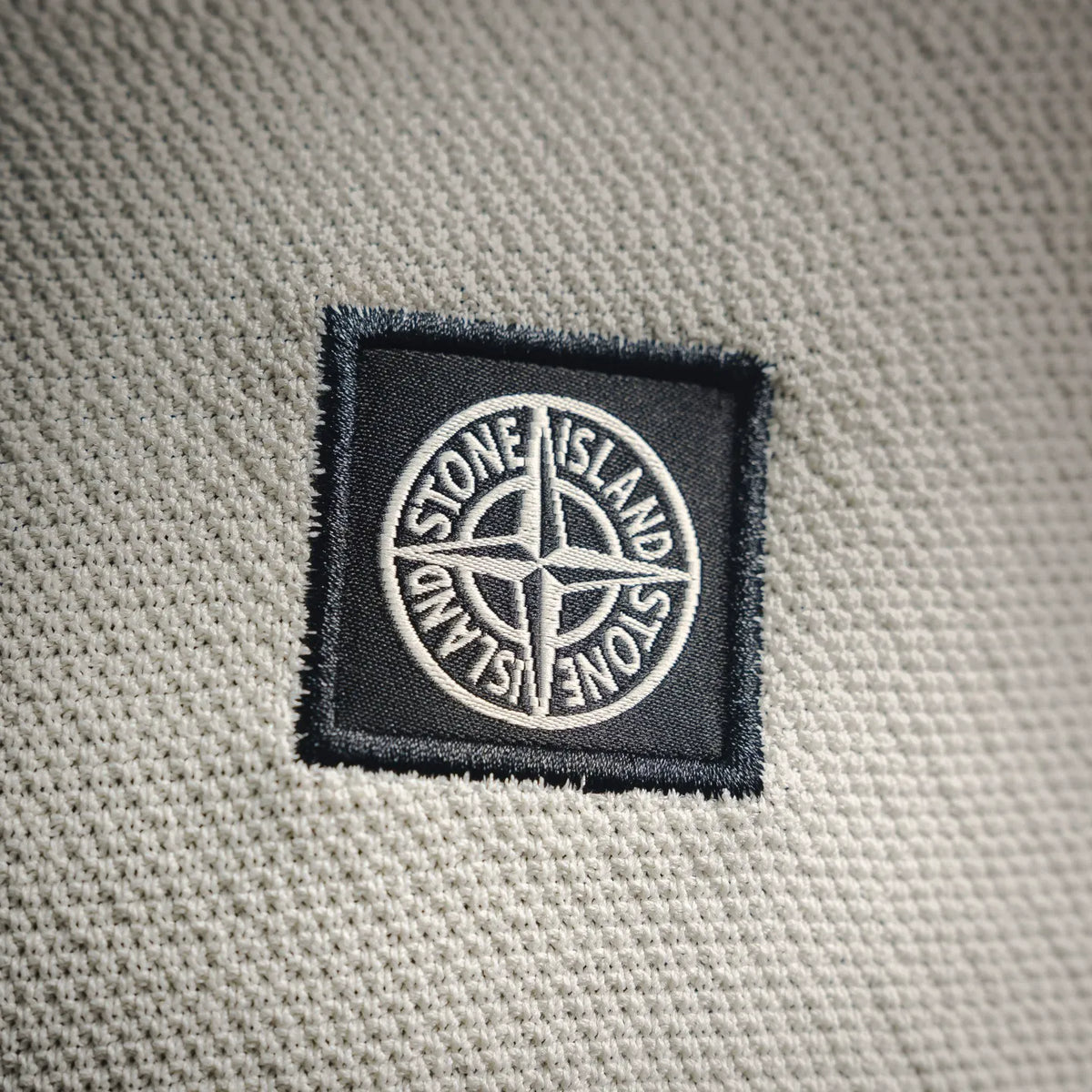 Stone Island logo patch on off-white polo shirt fabric, embroidered square label.