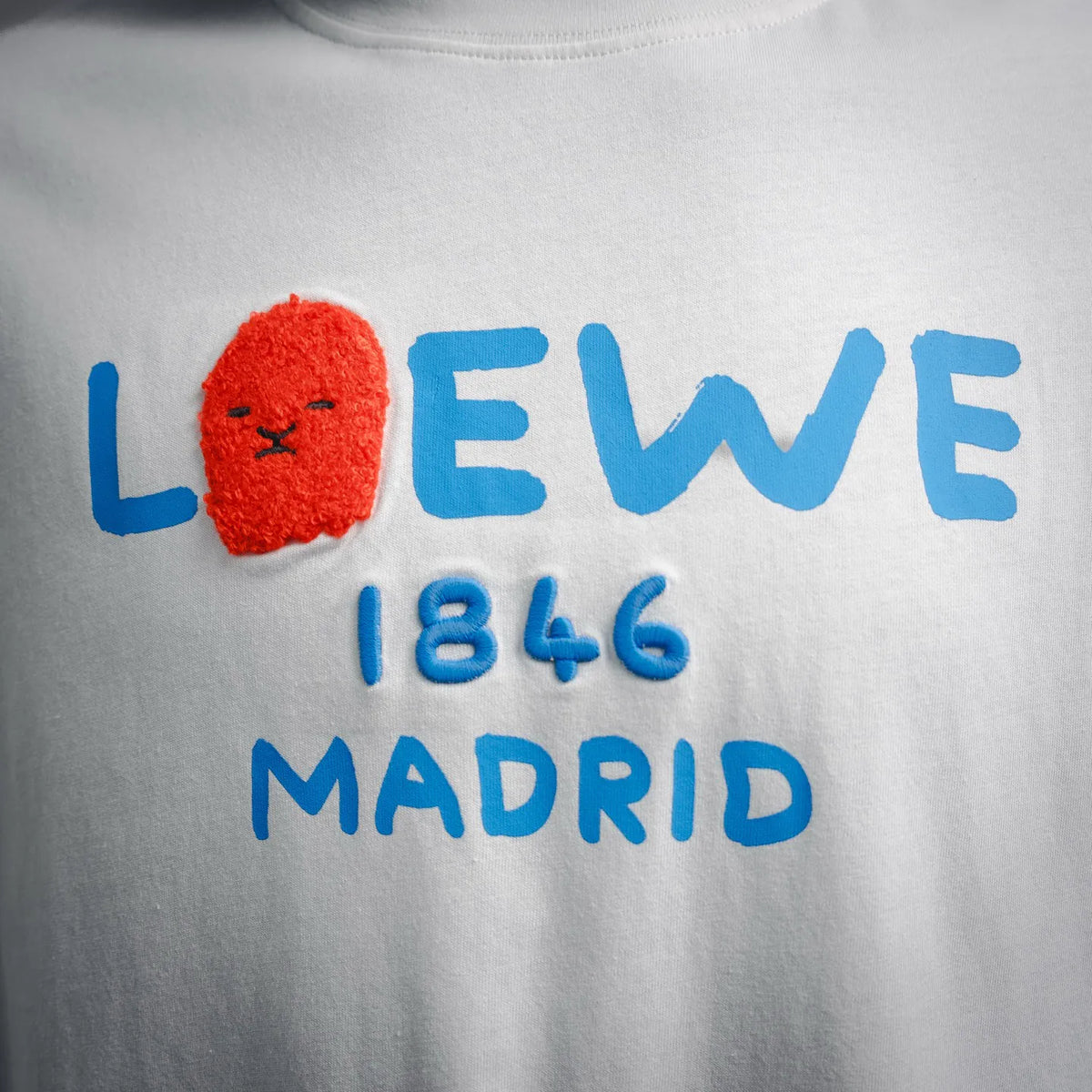 White LOEWE T-shirt front close-up: blue “LOEWE 1846 MADRID” text with red lion