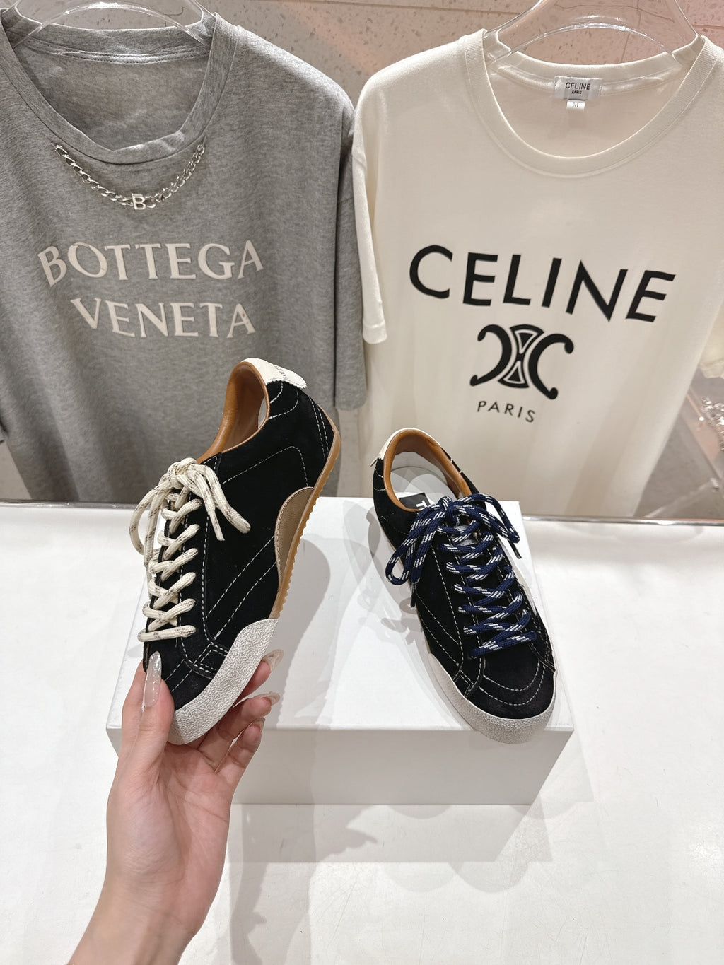 Hand holding a black and off-white low-top sneaker with white laces; pair on display.