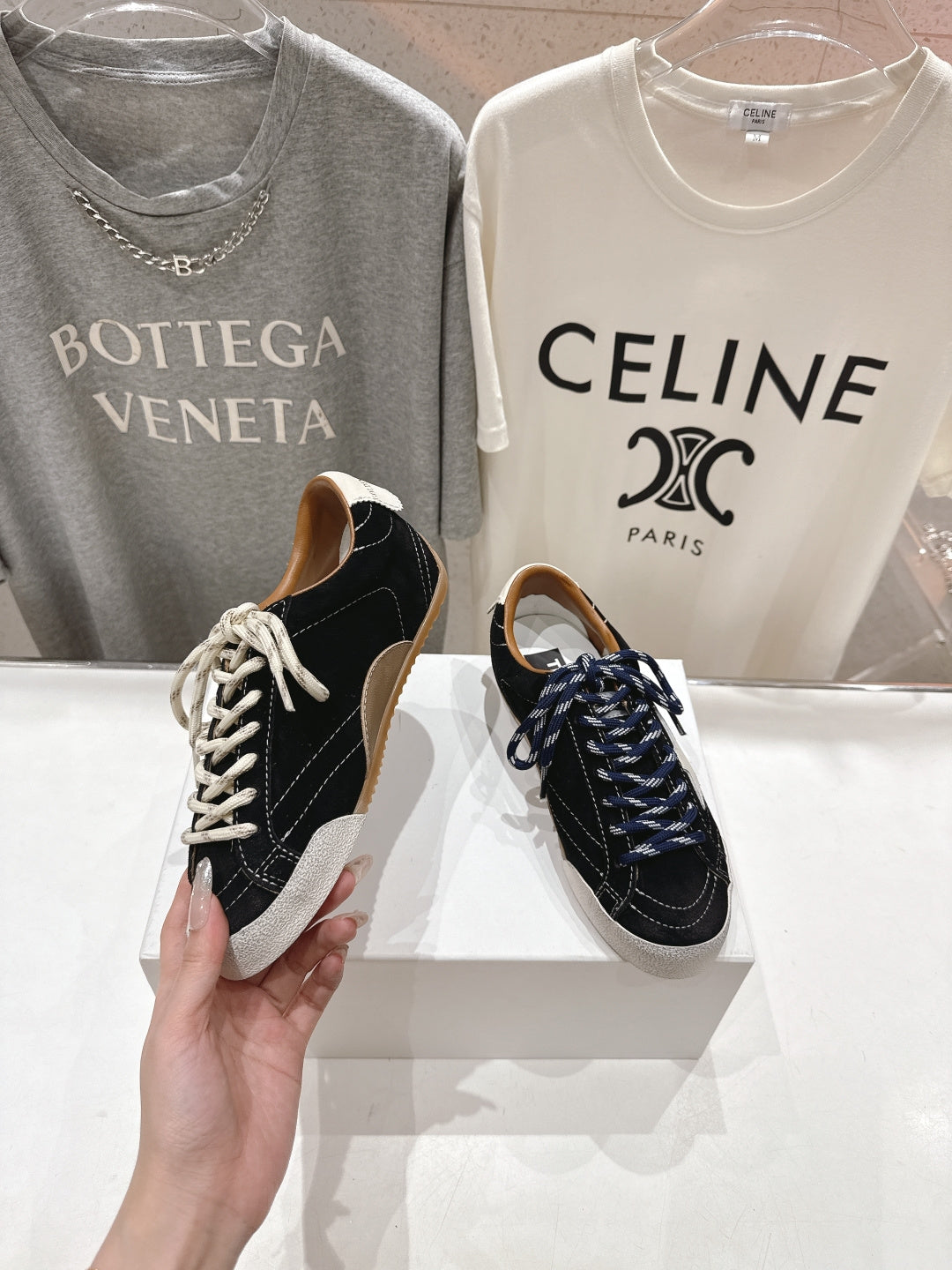 Hand holding a black and off-white low-top sneaker with white laces; pair on display.