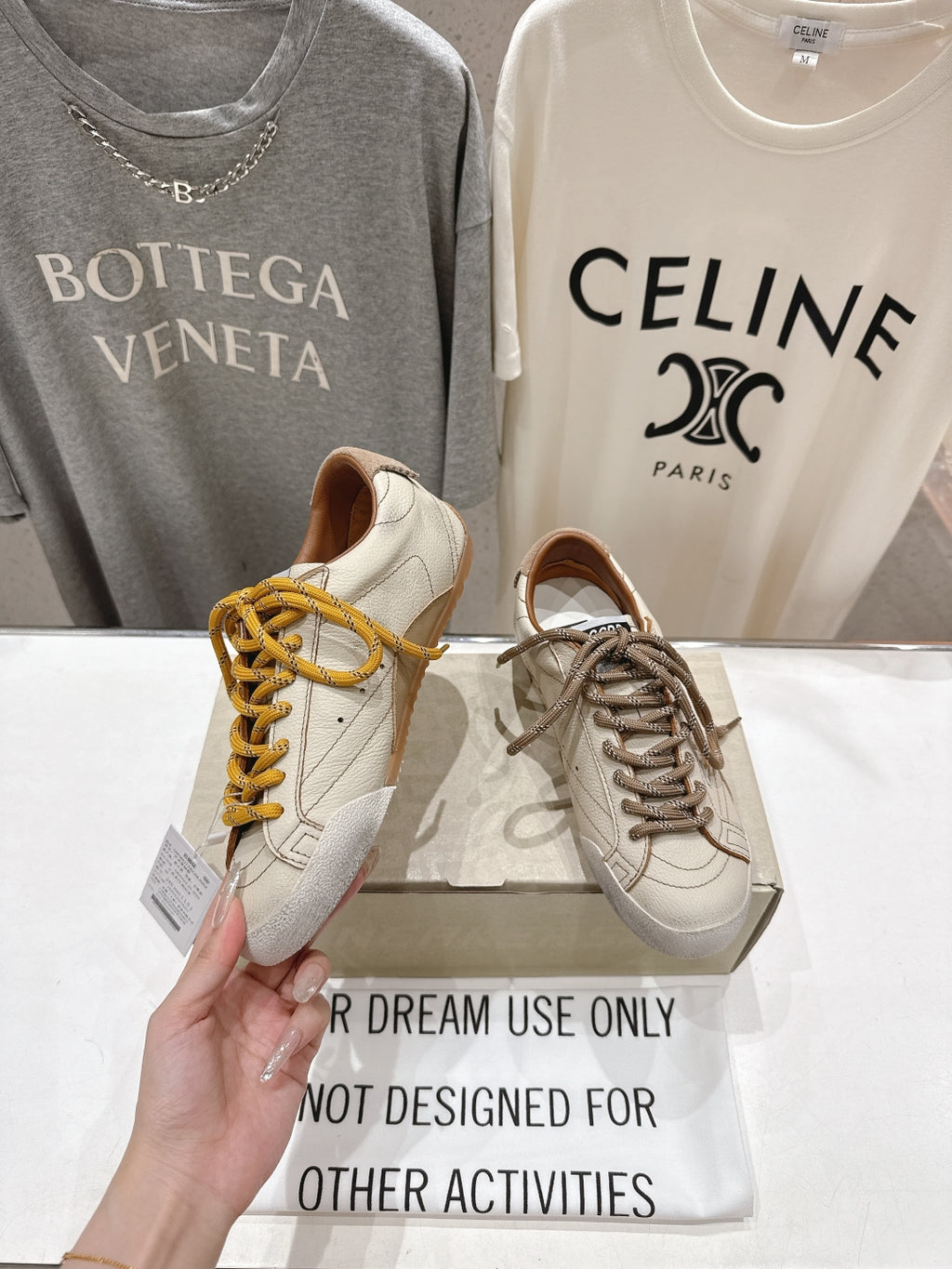 Golden Goose sneakers, hand holding white shoe with yellow laces beside brown-lace pair