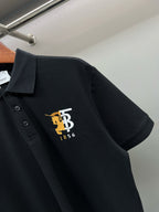 Black polo shirt with pointed collar and embroidered TB 1856 logo at left chest.
