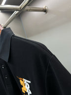 Side view of black polo shirt with embroidered TB 1856 logo at left chest.