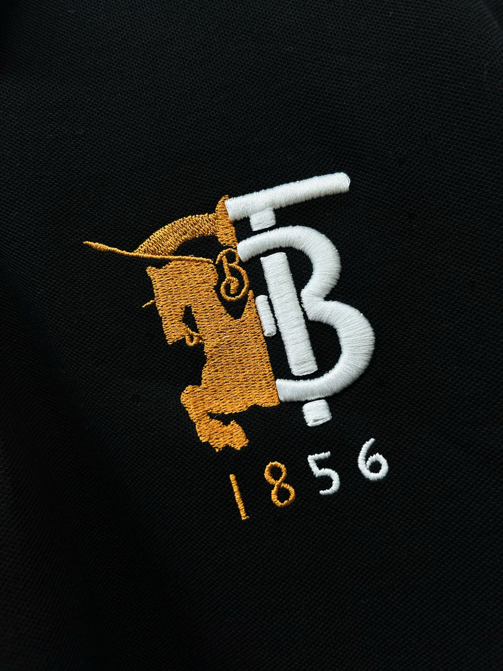 Close-up of embroidered TB 1856 logo on black polo shirt fabric.