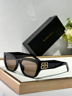 Balenciaga sunglasses with black frames, dark lenses, and gold B logo on arm.