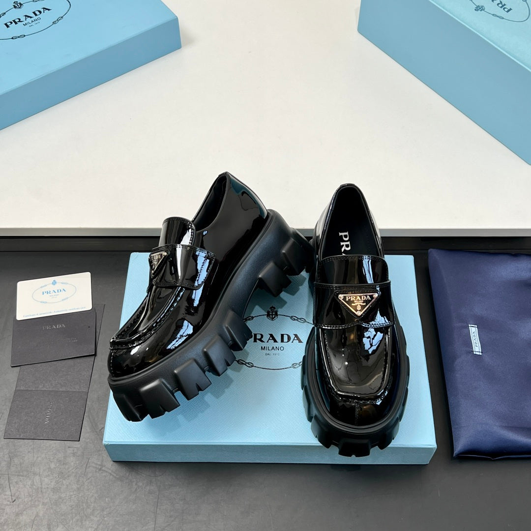 Two black patent loafers on blue box, showing strap buckle and lug platform soles